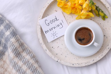 Cup of coffee, flowers and card with phrase GOOD MORNING! on bed, flat lay