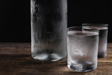 Bottle of vodka and shot glasses on wooden table against black background, closeup