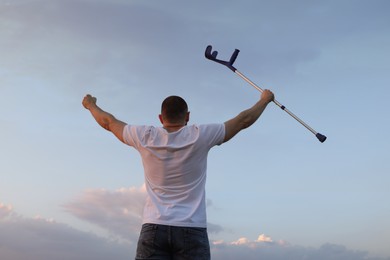 Man raising elbow crutch up to sky, back view. Healing miracle