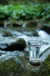 Glass of fresh water on stone near stream. Space for text