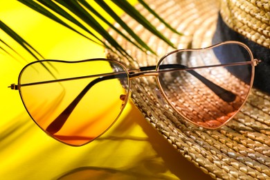 Stylish heart shaped sunglasses and straw hat on yellow background, closeup