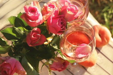 Photo of Glasses of delicious rose wine with petals, flowers and peaches on white picnic blanket outside, above view