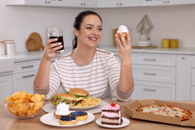 Happy overweight woman with glass of cola and cake in kitchen. Unhealthy food
