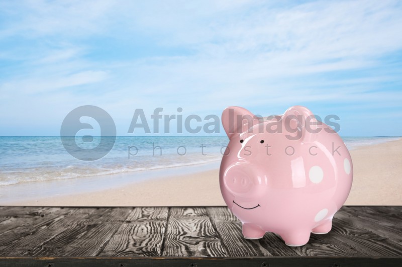 Saving money for summer vacation. Piggy bank on wooden surface near sandy beach and sea, space for text