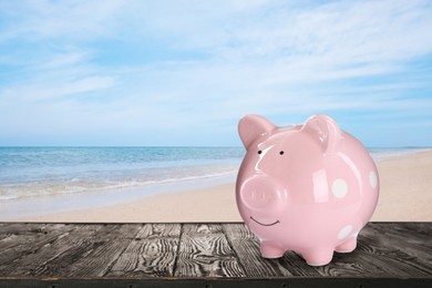 Saving money for summer vacation. Piggy bank on wooden surface near sandy beach and sea, space for text
