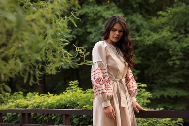 Beautiful woman wearing embroidered dress near wooden railing in countryside. Ukrainian national clothes