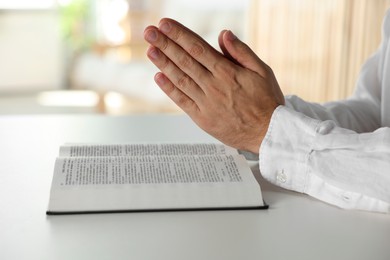 Man with Bible praying at white table indoors, closeup