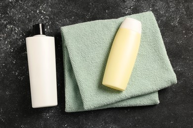Soft folded terry towel and cosmetic bottles on black textured background, flat lay