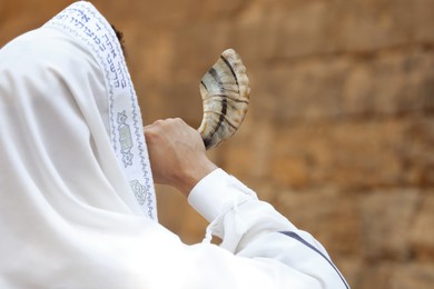 Jewish man blowing shofar on Rosh Hashanah outdoors. Wearing tallit with words Blessed Are You, Lord Our God, King Of The Universe, Who Has Sanctified Us With His Commandments, And Commanded Us To Enwrap Ourselves In Tzitziton in Hebrew