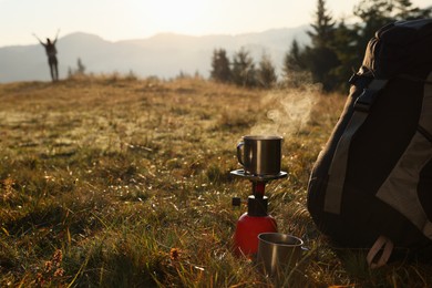 Camping burner with mug of hot drink near backpack on grass in mountains. Space for text
