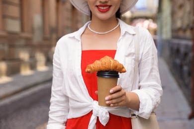 Woman holding tasty croissant and cup of coffee on city street, closeup