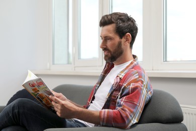 Smiling bearded man reading magazine on sofa indoors