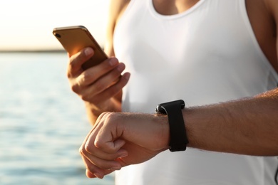 Man with fitness tracker and smartphone checking results after training near river, closeup