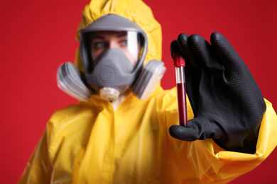 Woman in chemical protective suit holding test tube of blood sample against red background, focus on hand. Virus research