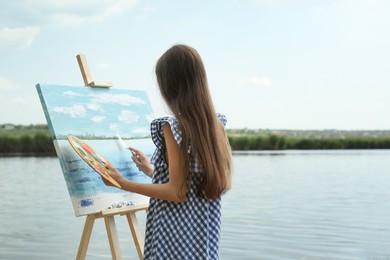 Little girl painting scenery on easel near lake, back view