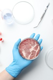Photo of Scientist holding Petri dish with cultured meat above white lab table, top view