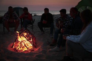 Group of friends gathering around bonfire on beach in evening. Camping season