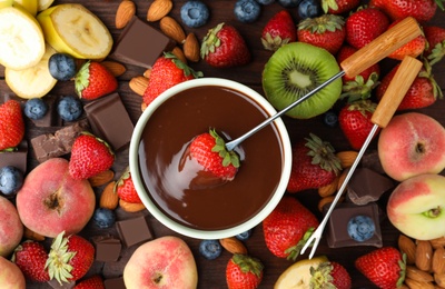 Fondue fork with strawberry in bowl of melted chocolate surrounded by other fruits on wooden table, flat lay