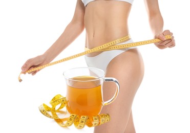 Slim young woman with measuring tape and glass cup of weight loss herbal tea on white background