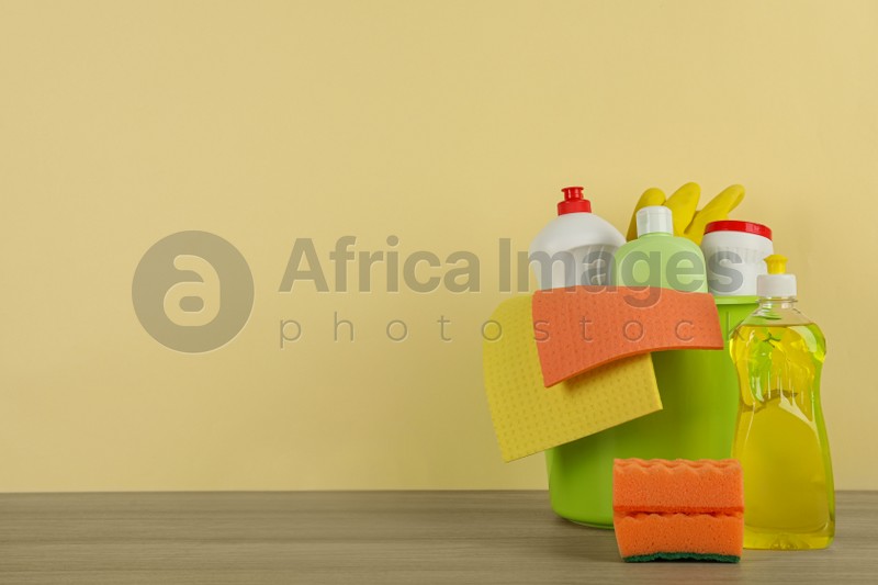 Bucket with different cleaning supplies on wooden floor near beige wall. Space for text