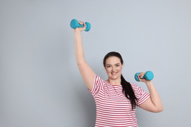 Happy overweight woman doing exercise with dumbbells on grey background, space for text