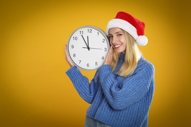 Woman in Santa hat with clock on yellow background, space for text. New Year countdown