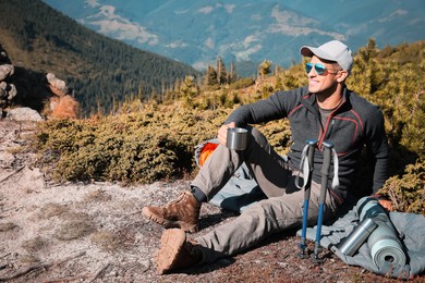 Hiker with mug of hot drink, trekking poles and other camping gear resting in mountains