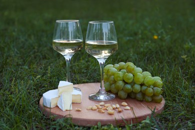 Photo of Two glasses of delicious white wine, grapes, cheese and nuts on green grass outdoors