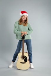 Young woman in Santa hat with acoustic guitar on light grey background. Christmas music