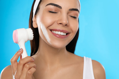 Young woman using facial cleansing brush on light blue background, closeup. Washing accessory