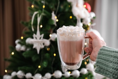 Woman with cup of delicious hot drink near Christmas tree at home, closeup. Space for text