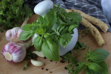 Photo of Mortar with different fresh herbs near garlic, horseradish roots and black peppercorns on wooden table, closeup