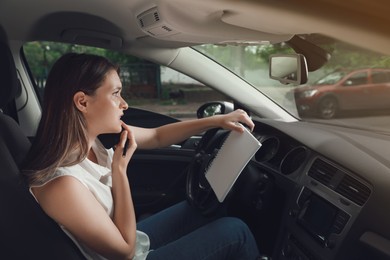 Stressed young woman with notebook talking on phone in driver's seat of modern car