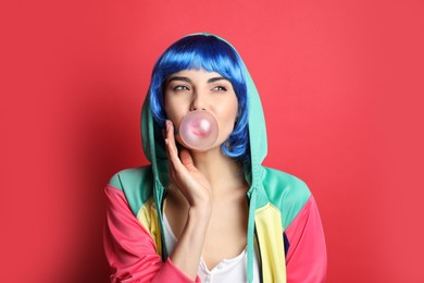 Fashionable young woman in colorful wig blowing bubblegum on red background
