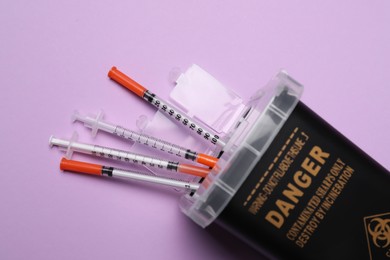 Disposable syringes and sharps container on violet background, top view