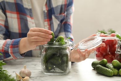 Woman putting cucumber into glass jar at white marble kitchen table, closeup. Pickling vegetables