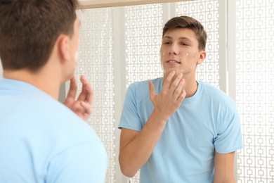 Photo of Teen guy with acne problem applying cream near mirror indoors