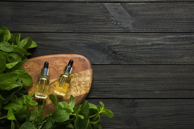 Bottles of mint essential oil and green leaves on black wooden table, flat lay. Space for text