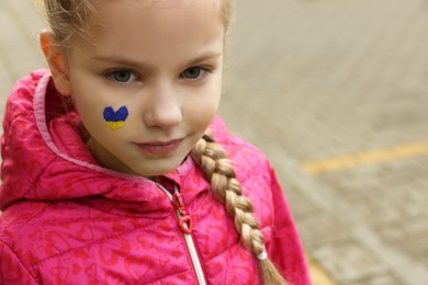 Photo of Little girl with drawing of Ukrainian flag on face in heart shape outdoors, space for text