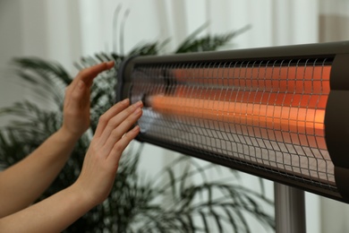 Woman warming hands near electric infrared heater indoors, closeup