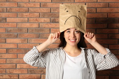 Happy woman wearing paper bag with drawn sad face near brick wall