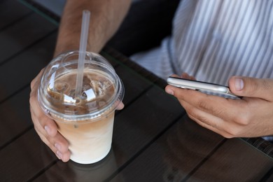 Man with smartphone and plastic takeaway cup of delicious iced coffee at table in cafe, closeup