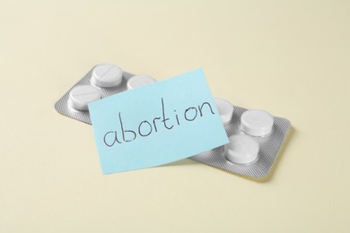 Paper note with word Abortion and pills on beige background, closeup