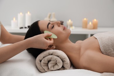 Photo of Young woman receiving facial massage with gua sha tool in beauty salon