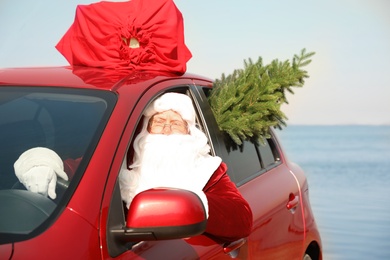 Photo of Authentic Santa Claus with fir tree and bag full of presents on roof driving modern car  near sea