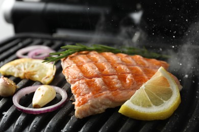 Tasty salmon cooking with rosemary, garlic, onion and lemon on electric grill, closeup