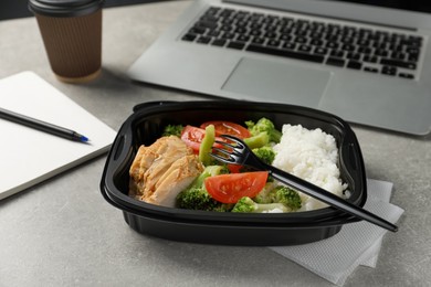 Photo of Container with tasty food, laptop, fork and notebook on light grey table. Business lunch