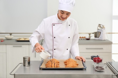 Photo of Young female pastry chef pouring chocolate sauce onto croissants at table in kitchen