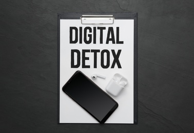 Clipboard with phrase DIGITAL DETOX, smartphone and earphones on black stone table, flat lay