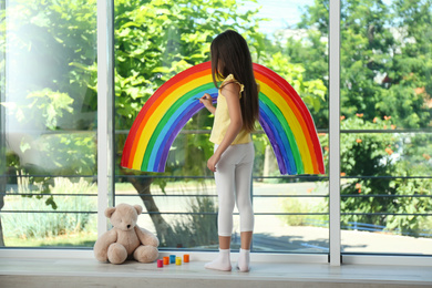 Little girl drawing rainbow on window with paints indoors. Stay at home concept